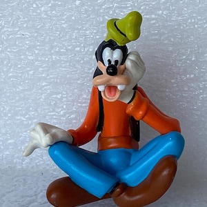 以下が含まれることがあります： ディズニーの漫画のキャラクター、グーフィーのカラフルなフィギュア。座ったポーズで、オレンジ色のシャツ、青いズボン、茶色の靴、緑の帽子を着用しています。口を開けて、顔の近くに手を置いています。