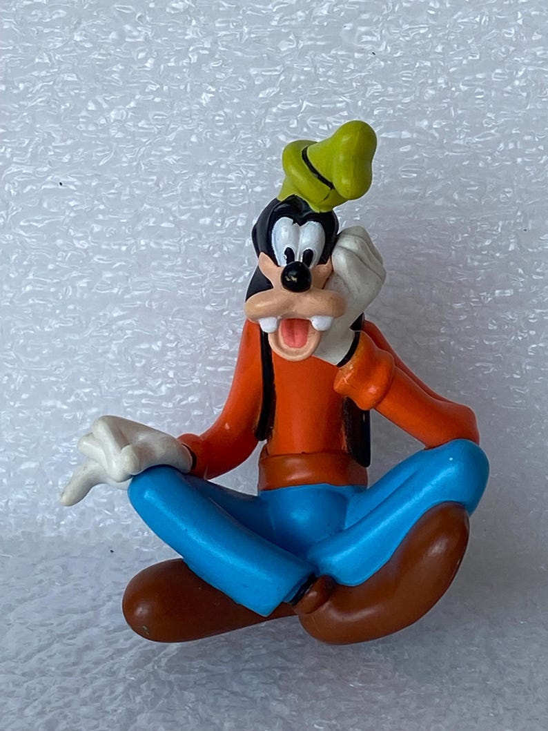 以下が含まれることがあります： ディズニーの漫画のキャラクター、グーフィーのカラフルなフィギュア。座ったポーズで、オレンジ色のシャツ、青いズボン、茶色の靴、緑の帽子を着用しています。口を開けて、顔の近くに手を置いています。