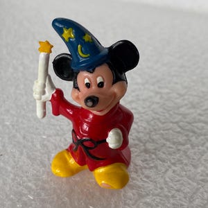 Pu&ograve; includere: Una piccola statuetta in plastica di Topolino vestito da stregone, che indossa un cappello a punta blu con stelle gialle e una mezzaluna, una tunica rossa con cintura nera e scarpe gialle, mentre tiene in mano una bacchetta bianca.