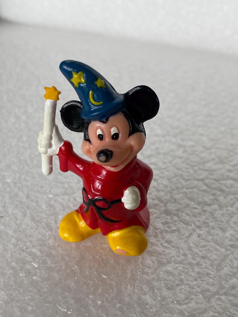 Pu&ograve; includere: Una piccola statuetta in plastica di Topolino vestito da stregone, che indossa un cappello a punta blu con stelle gialle e una mezzaluna, una tunica rossa con cintura nera e scarpe gialle, mentre tiene in mano una bacchetta bianca.