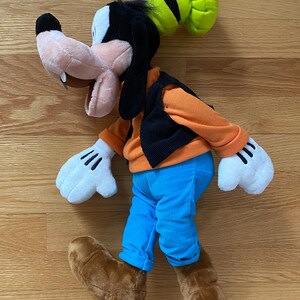 Może przedstawiać: Pluszowa zabawka przedstawiająca postać Goofy'ego z brzoskwiniowym pyszczkiem, czarnymi uszami i zielonym kapeluszem. Figurka ma na sobie pomarańczową bluzkę z długim rękawem, czarną kamizelkę, jasnoniebieskie spodnie, białe rękawiczki i brązowe buty.