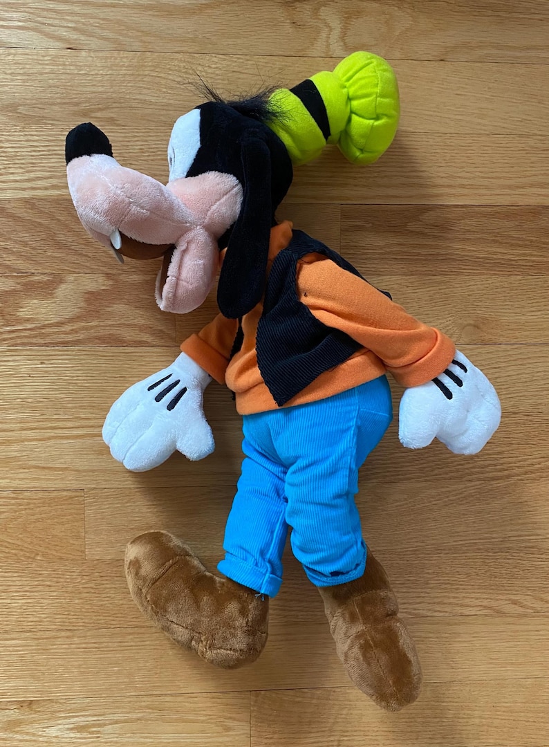Może przedstawiać: Pluszowa zabawka przedstawiająca postać Goofy'ego z brzoskwiniowym pyszczkiem, czarnymi uszami i zielonym kapeluszem. Figurka ma na sobie pomarańczową bluzkę z długim rękawem, czarną kamizelkę, jasnoniebieskie spodnie, białe rękawiczki i brązowe buty.