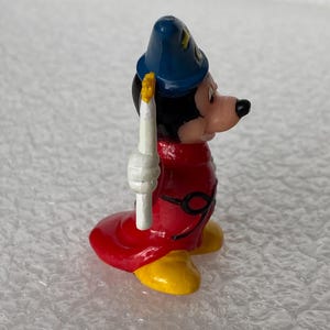 Pu&ograve; includere: Una piccola statuetta di un personaggio che indossa un cappello a punta blu con dettagli a stella gialli, una lunga veste rossa con una cintura nera e scarpe gialle. Il personaggio ha la testa nera, il viso color carne e tiene una candela bianca.