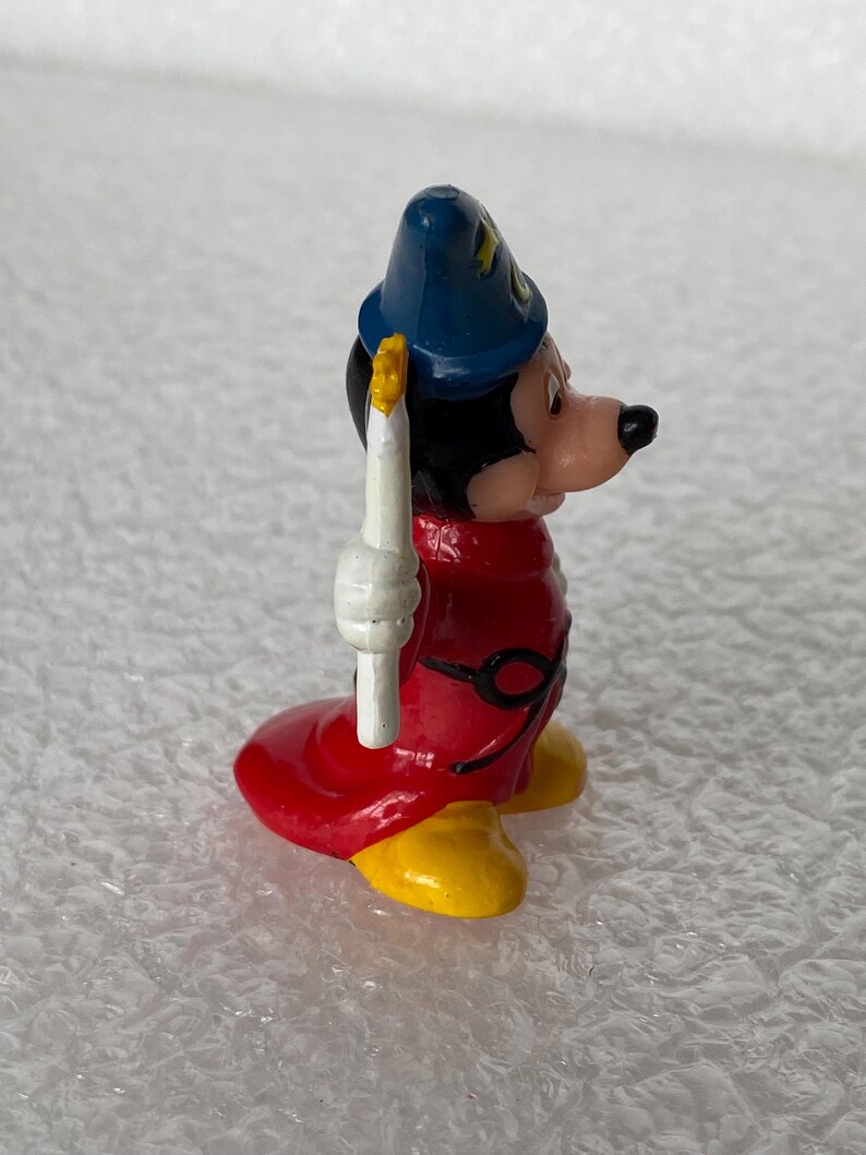 Pu&ograve; includere: Una piccola statuetta di un personaggio che indossa un cappello a punta blu con dettagli a stella gialli, una lunga veste rossa con una cintura nera e scarpe gialle. Il personaggio ha la testa nera, il viso color carne e tiene una candela bianca.