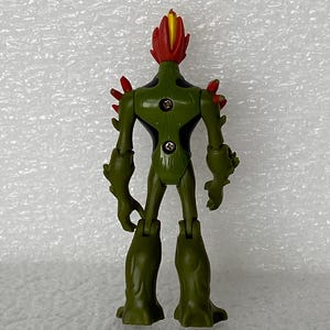 May include: A green humanoid action figure with a red and yellow flame-like head, red spikes on the shoulders, and black accents on the torso. The back of the figure features two visible metal screws and articulated joints at the shoulders and hips.