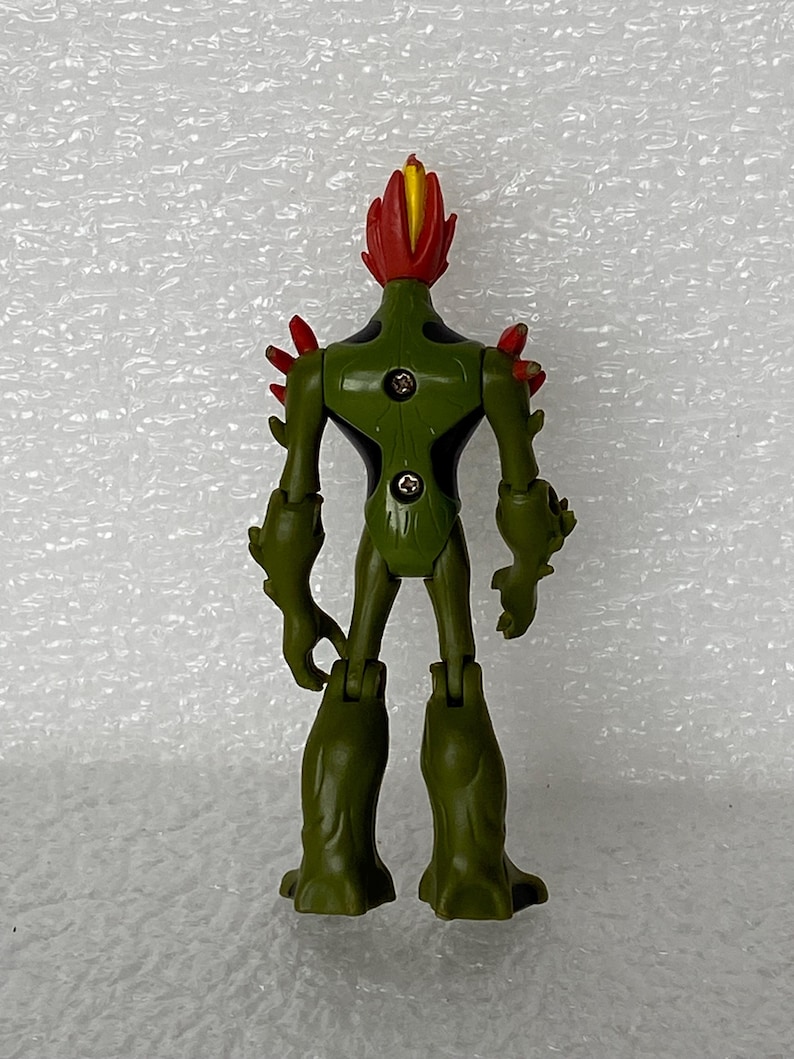 May include: A green humanoid action figure with a red and yellow flame-like head, red spikes on the shoulders, and black accents on the torso. The back of the figure features two visible metal screws and articulated joints at the shoulders and hips.