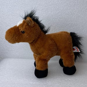 May include: A plush toy horse in a standing position. The horse has a brown body, black mane and tail, and black hooves. It has a white patch on its forehead.