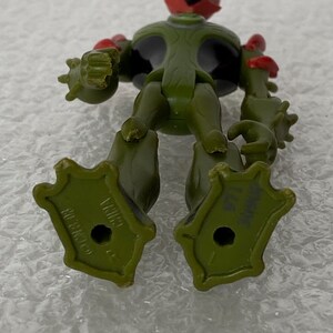May include: An olive green action figure with a black chest, red head accents, and a yellow nose. The figure has textured arms, a wide stance, and large feet with holes in the center. The soles are marked with the text CN & 08 and CHINA.