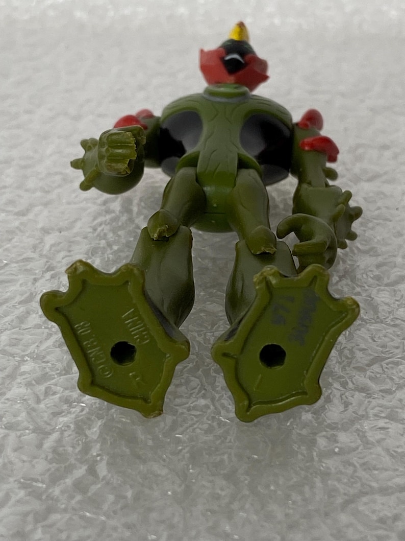 May include: An olive green action figure with a black chest, red head accents, and a yellow nose. The figure has textured arms, a wide stance, and large feet with holes in the center. The soles are marked with the text CN & 08 and CHINA.