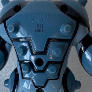 May include: A blue and black toy figure with the text "OCN B.10 CHINA 971 80010" printed on the back. The toy has a rounded shape with black accents and protruding elements.