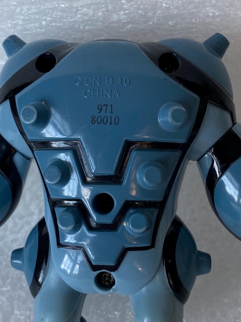 May include: A blue and black toy figure with the text "OCN B.10 CHINA 971 80010" printed on the back. The toy has a rounded shape with black accents and protruding elements.