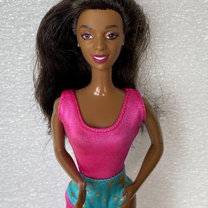 May include: A doll with dark brown hair, brown skin, and painted facial features including pink eyeshadow and bright pink lipstick. The doll wears a sleeveless hot pink top and a teal blue bottom piece featuring a small gold star pattern.