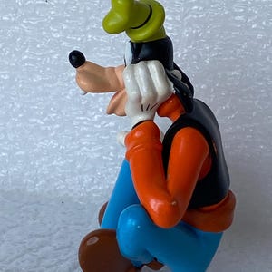 以下が含まれることがあります： ディズニーの漫画のキャラクター、グーフィーのフィギュア。座った姿勢で、黒いベスト、オレンジ色の袖、青いズボン、緑の帽子を着用しています。手は白く、鼻はタン色です。