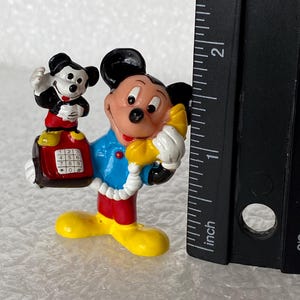 May include: A Mickey Mouse figurine wearing a blue shirt and red pants, holding a yellow telephone receiver to an ear. A smaller Mickey Mouse figure sits on a red telephone base with a keypad. A black ruler shows the height is under 2 inches.
