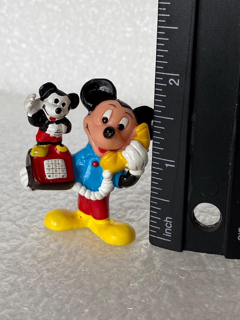 May include: A Mickey Mouse figurine wearing a blue shirt and red pants, holding a yellow telephone receiver to an ear. A smaller Mickey Mouse figure sits on a red telephone base with a keypad. A black ruler shows the height is under 2 inches.