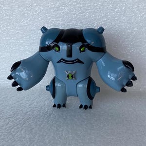 May include: A light blue action figure with black accents, featuring a stylized design. The figure has large arms and feet with black claws, green eyes, and a symbol on its chest. The figure is a toy.