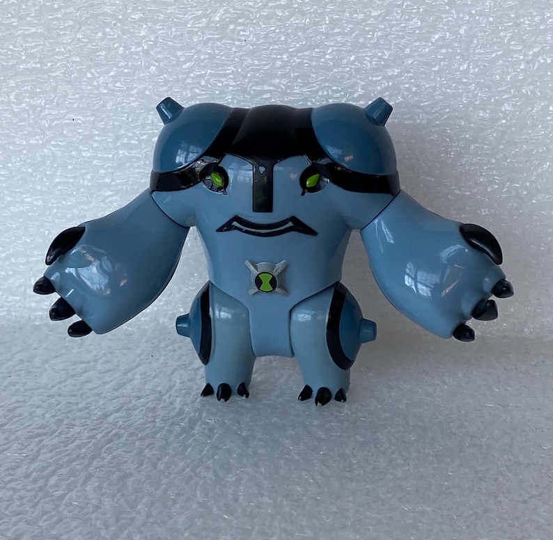 May include: A light blue action figure with black accents, featuring a stylized design. The figure has large arms and feet with black claws, green eyes, and a symbol on its chest. The figure is a toy.