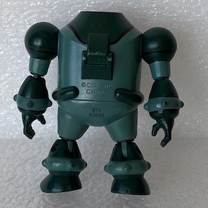May include: A dark green toy robot with articulated limbs and a rounded body. The robot has a head, two arms, and two legs. The text "CN B 10 CHINA" and "971 51010" are printed on the robot's body. The toy is made of plastic.
