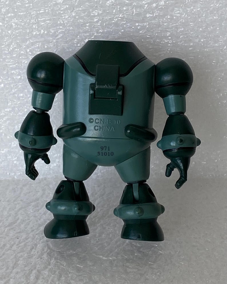 May include: A dark green toy robot with articulated limbs and a rounded body. The robot has a head, two arms, and two legs. The text "CN B 10 CHINA" and "971 51010" are printed on the robot's body. The toy is made of plastic.
