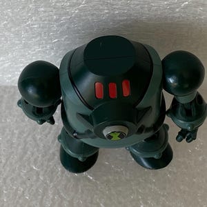 May include: A green and black action figure with a rounded head, two large arm-like spheres, and two leg-like spheres. The figure has three red buttons on its chest and a green and black emblem on its abdomen.