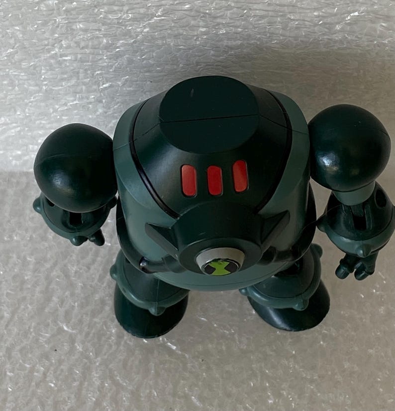 May include: A green and black action figure with a rounded head, two large arm-like spheres, and two leg-like spheres. The figure has three red buttons on its chest and a green and black emblem on its abdomen.