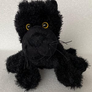 May include: A plush black panther toy with yellow eyes and whiskers. The stuffed animal is made of soft, fuzzy material and has a friendly expression. The panther is lying down, with its paws extended.