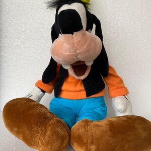 Może przedstawiać: Pluszowa zabawka przedstawiająca postać Goofy'ego w zielonym kapeluszu, z czarnymi uszami, brzoskwiniowym pyszczkiem i dwoma białymi zębami. Postać ma na sobie pomarańczową koszulkę z długim rękawem, czarną kamizelkę, niebieskie spodnie, białe rękawiczki.