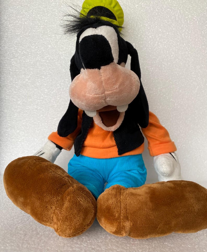 Może przedstawiać: Pluszowa zabawka przedstawiająca postać Goofy'ego w zielonym kapeluszu, z czarnymi uszami, brzoskwiniowym pyszczkiem i dwoma białymi zębami. Postać ma na sobie pomarańczową koszulkę z długim rękawem, czarną kamizelkę, niebieskie spodnie, białe rękawiczki.