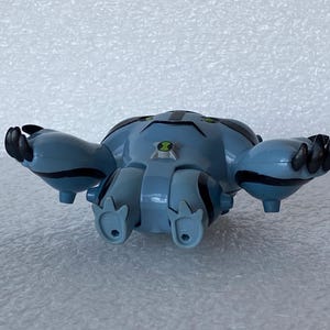 May include: A blue and black toy figure of a character with a round body, four legs, and two arms. The figure has black claws and green accents. A green and black emblem is on the chest.