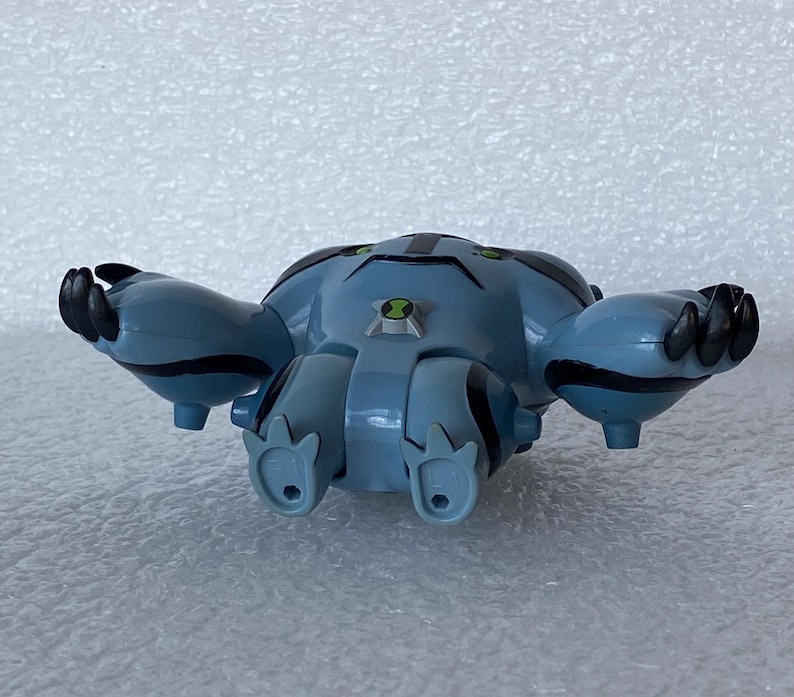 May include: A blue and black toy figure of a character with a round body, four legs, and two arms. The figure has black claws and green accents. A green and black emblem is on the chest.