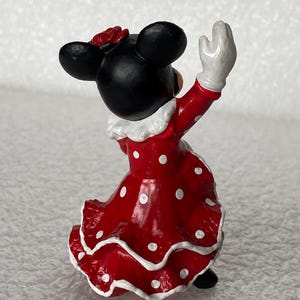 May include: A small figurine of a character with black ears wearing a red dress with white polka dots and a ruffled hem. The character has a white collar, white gloves, and a red flower accessory on the head, shown from a rear-facing perspective.