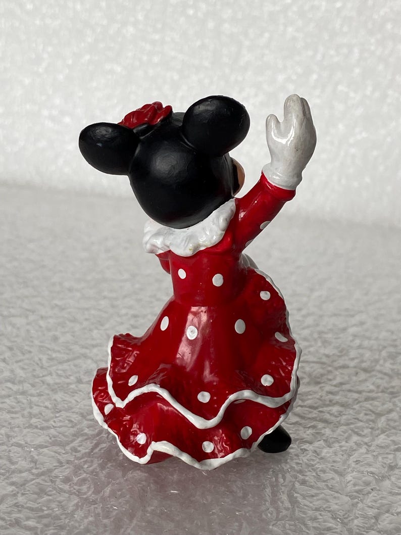 May include: A small figurine of a character with black ears wearing a red dress with white polka dots and a ruffled hem. The character has a white collar, white gloves, and a red flower accessory on the head, shown from a rear-facing perspective.