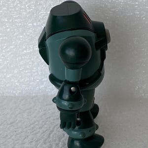 May include: A dark green robot toy with a rounded head, spherical shoulder and knee joints, and a cylindrical body. The robot has a small red accent on its head. It is standing on a white surface.