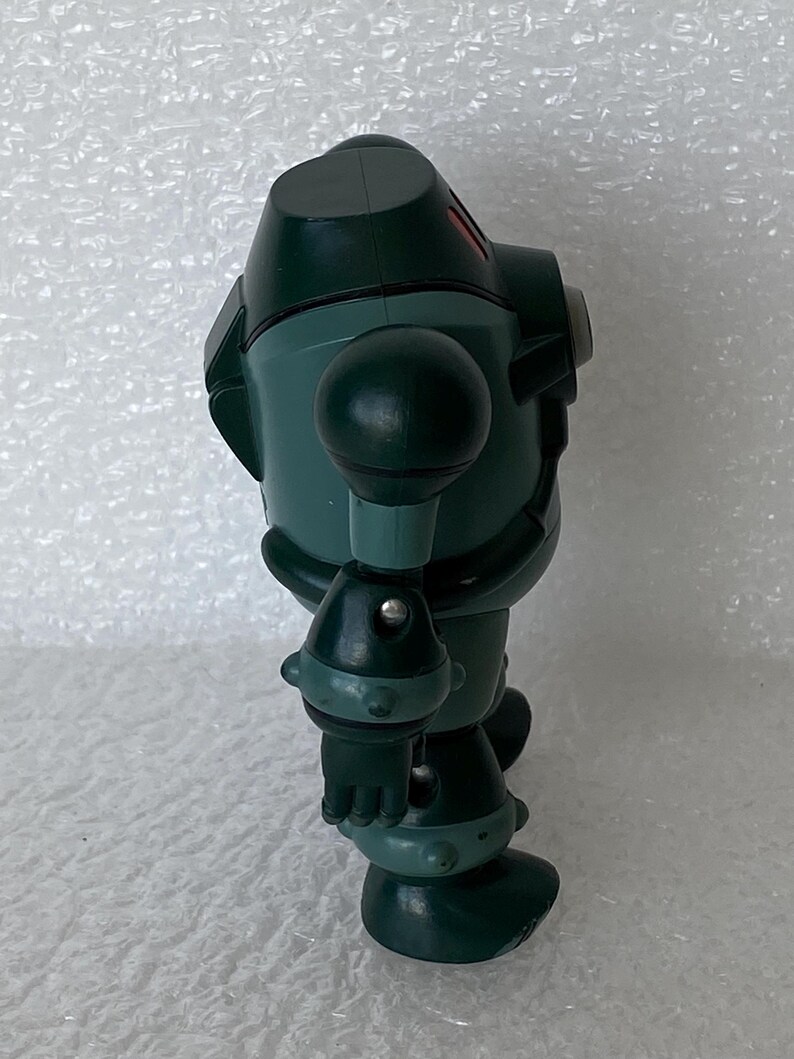 May include: A dark green robot toy with a rounded head, spherical shoulder and knee joints, and a cylindrical body. The robot has a small red accent on its head. It is standing on a white surface.