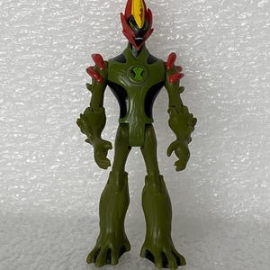 May include: A Swampfire action figure from Ben 10 featuring a green plant-like body with black accents, red shoulder spikes, and a flame-shaped head with a yellow crest. The chest displays a green circular emblem with a black hourglass-shaped symbol.