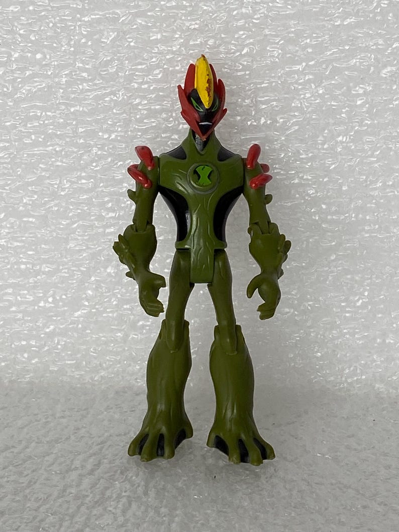 May include: A Swampfire action figure from Ben 10 featuring a green plant-like body with black accents, red shoulder spikes, and a flame-shaped head with a yellow crest. The chest displays a green circular emblem with a black hourglass-shaped symbol.