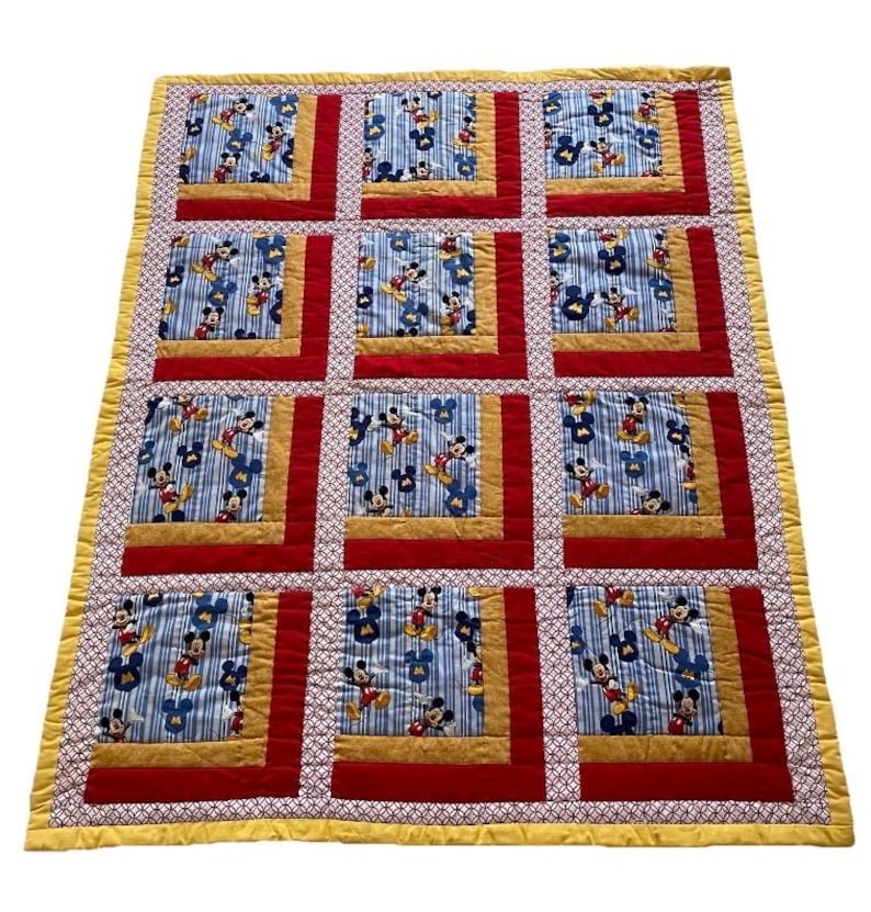 Pode incluir: Um edredom feito &agrave; m&atilde;o com um padr&atilde;o repetido do Mickey Mouse sobre um fundo listrado azul. O edredom tem bordas vermelhas e douradas e uma borda amarela estampada. O edredom &eacute; provavelmente para a cama ou o quarto de uma crian&ccedil;a.