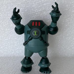May include: A teal action figure with a circular emblem on its chest and three red lights on its head. The figure has articulated arms and legs, with black accents and a green symbol. The figure is standing on a white surface.