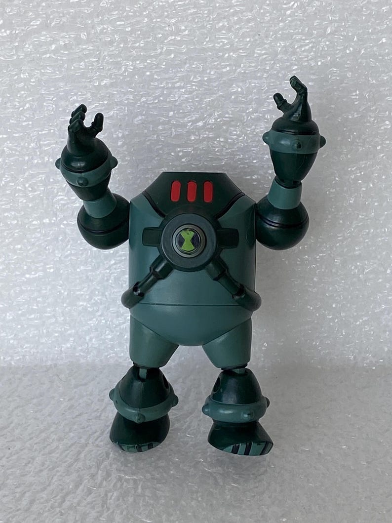 May include: A teal action figure with a circular emblem on its chest and three red lights on its head. The figure has articulated arms and legs, with black accents and a green symbol. The figure is standing on a white surface.