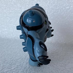 May include: A blue and black action figure of a fictional character with a rounded head, spiky protrusions, and claw-like hands. The figure has black accents and green eyes. The toy is a collectible item.