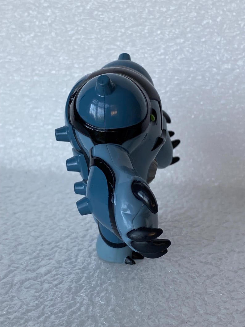 May include: A blue and black action figure of a fictional character with a rounded head, spiky protrusions, and claw-like hands. The figure has black accents and green eyes. The toy is a collectible item.