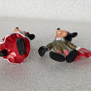 May include: Two small Disney figurines featuring Mickey and Minnie Mouse in matador-inspired outfits. The figures are shown from an overhead angle, revealing black shoes with the text Disney and Made in Germany molded into the soles.
