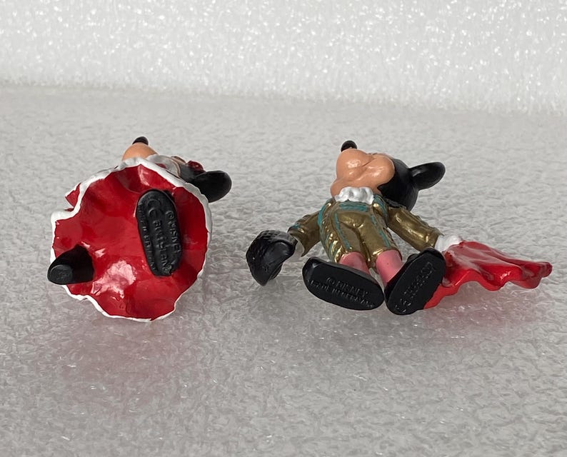 May include: Two small Disney figurines featuring Mickey and Minnie Mouse in matador-inspired outfits. The figures are shown from an overhead angle, revealing black shoes with the text Disney and Made in Germany molded into the soles.