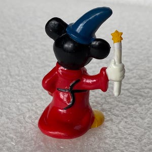 Pu&ograve; includere: Una piccola statuetta di un personaggio con orecchie nere che indossa un cappello a punta blu e una tunica rossa. Il personaggio ha mani guantate di bianco e tiene una bacchetta bianca sormontata da una stella gialla, vista da dietro.