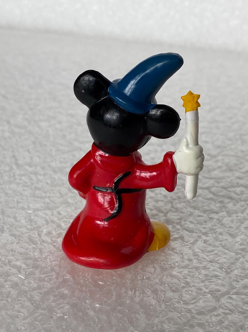 Pu&ograve; includere: Una piccola statuetta di un personaggio con orecchie nere che indossa un cappello a punta blu e una tunica rossa. Il personaggio ha mani guantate di bianco e tiene una bacchetta bianca sormontata da una stella gialla, vista da dietro.