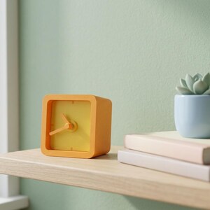 Può includere: Un orologio analogico quadrato con cornice arancione e quadrante giallo con lancette arancioni. Accanto all'orologio ci sono due libri impilati e un piccolo vaso azzurro contenente una pianta succulenta verde, su una mensola in legno chiaro.