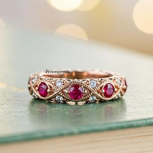 May include: Rose gold ring featuring a repeating pattern of red gemstones and small clear stones. The ring is set on a green book, with a blurred background of soft, out-of-focus lights.