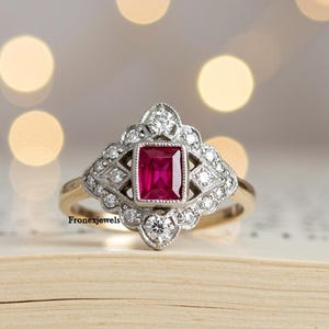 Peut inclure: Une bague de style vintage avec une pierre rouge foncé rectangulaire au centre, entourée d'un motif géométrique orné de diamants. La bague a un anneau en or et se détache sur un fond flou.