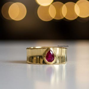 May include: A gold ring featuring a teardrop-shaped, deep red gemstone. The ring has a wide band and a polished finish. The gemstone is set in a bezel setting. The ring is displayed on a white surface.