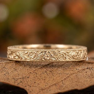 May include: A gold-colored ring with an intricate floral pattern. The ring is placed on a brown leaf, with a blurred background of fall colors. The ring's design features detailed carvings of flowers and leaves, creating a vintage or antique aesthetic.